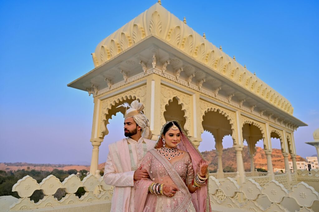This image shows a couple in traditional wedding attire standing in front of a white, intricately carved architectural structure reminiscent of a palace pavilion. | Jaipur Wedding Guide