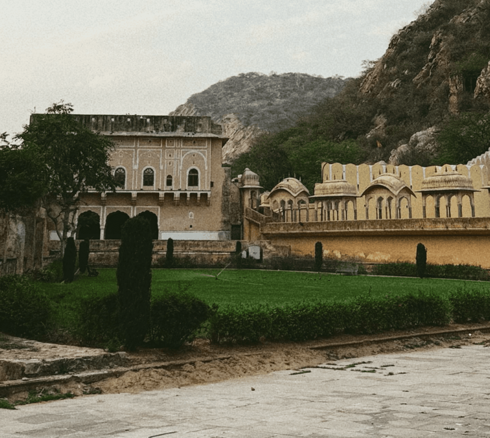 Painted palace facade and domed pavilions of Sisodia Rani Garden beside a green lawn with cypress trees against rocky Aravalli hills in Jaipur