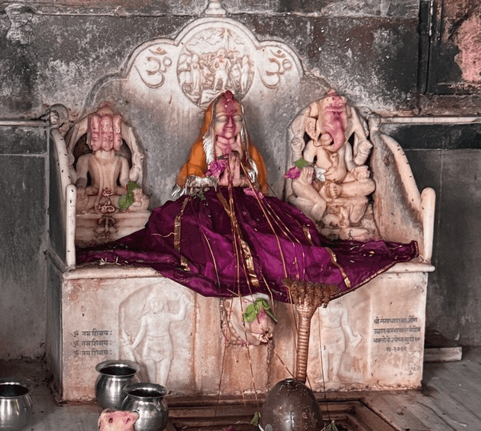 Marble shrine at Galtaji Temple in Jaipur with three carved deities, the central idol draped in purple cloth beneath carved Om symbols