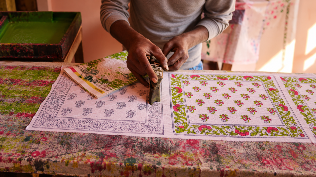 Artisan pressing a carved wooden block onto cotton fabric to print pink and green floral patterns in Jaipur